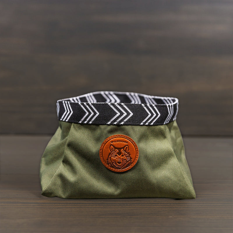 The Backpacking Bowl is a small olive green fabric bag with a chevron black-and-white rim and a brown leather wolf patch—ideal as a portable or travel dog water bowl, shown on a wooden background.