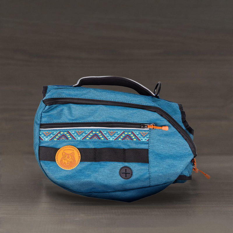 The Dog Backpack is a blue fabric waist bag with orange zippers, tribal pattern, circular orange patch, and black handle. Durable and water resistant, it’s perfect for hiking dog gear and shown on a plain brown background.