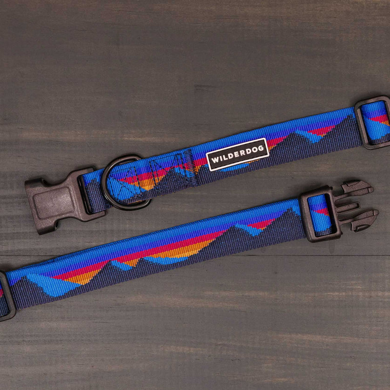 Two blue Cordillera Ultralight Collars with red and orange mountain patterns are displayed on a dark wooden surface. These collars feature strong nylon webbing, heavy-duty plastic clips, and adjustable straps.