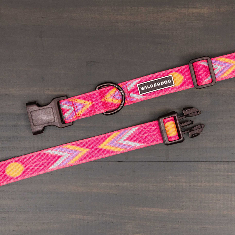 The Playa Ultralight Collar features a pink nylon webbing with orange and gray arrow patterns, a sturdy plastic clip, metal D-ring, and "Wilderdog" label. It's shown on a dark wooden surface.