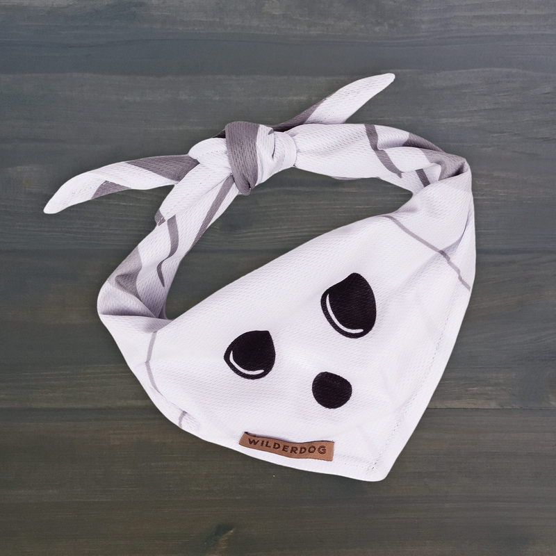 Ghost Bandana—white, quick-dry, black ghost face, brown Wilderdog tag—tied on a dark wood surface.