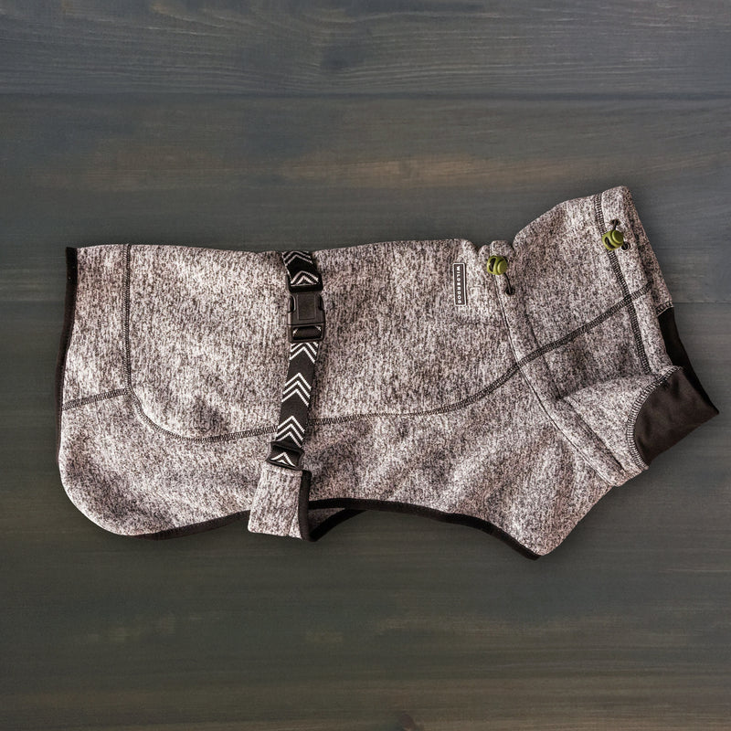 A Fleece Jacket for dogs in gray and black, featuring textured fabric, green cord locks, and an adjustable strap, is shown laid flat on a dark wooden surface.