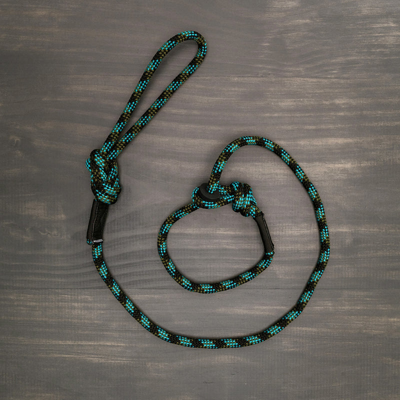 A Granite Slip Lead, with its black and teal woven design reminiscent of a rock climbing rope, is laid out on a wooden surface. The slip lead features two loops: a larger one at one end and another smaller loop formed in the middle with a knot. The texture of the wooden background accentuates this durable slip lead's details.