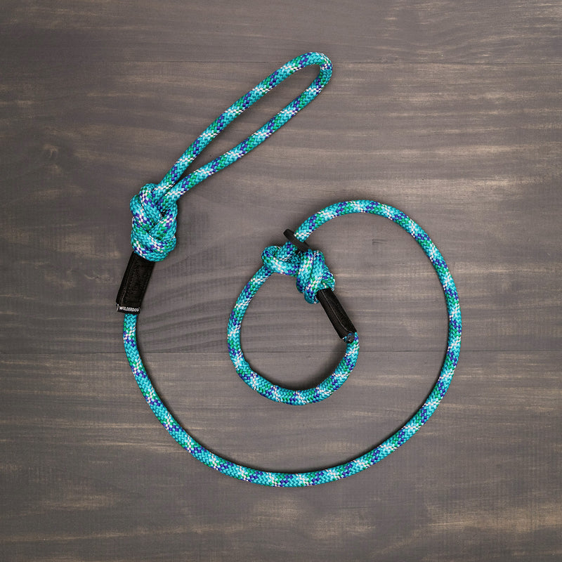 The Islander Reflective Slip Lead, a blue and green patterned dog leash made from durable rock climbing rope, features two knots and black leather accents. It is neatly arranged in a circular shape on a dark wooden surface, showcasing its sturdiness and stylish design.