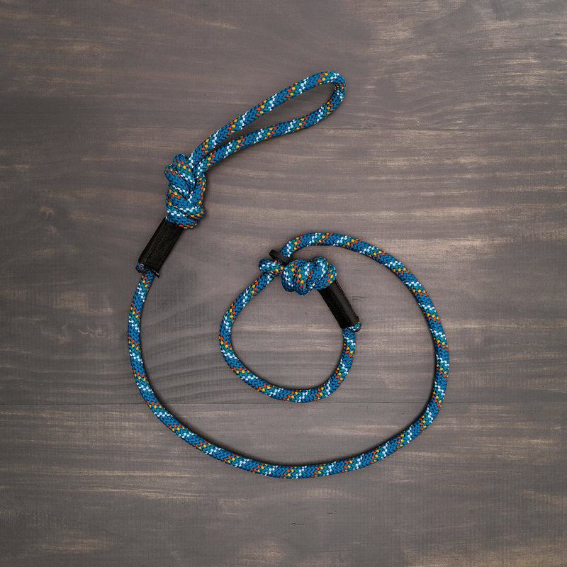 The neatly coiled Pacific Blue Reflective Slip Lead, featuring a braided pattern in blue and multicolor, lies on a dark wooden surface. This durable dog leash with a loop handle and metal clip is made from rock climbing rope slip lead material, ensuring it is both sturdy and perfect for long-lasting use.