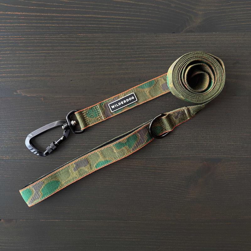 The Camo Ultralight Leash, featuring green camouflage-patterned nylon, a black metal clip, and a "Wild One" label, is neatly coiled on a dark wooden surface.
