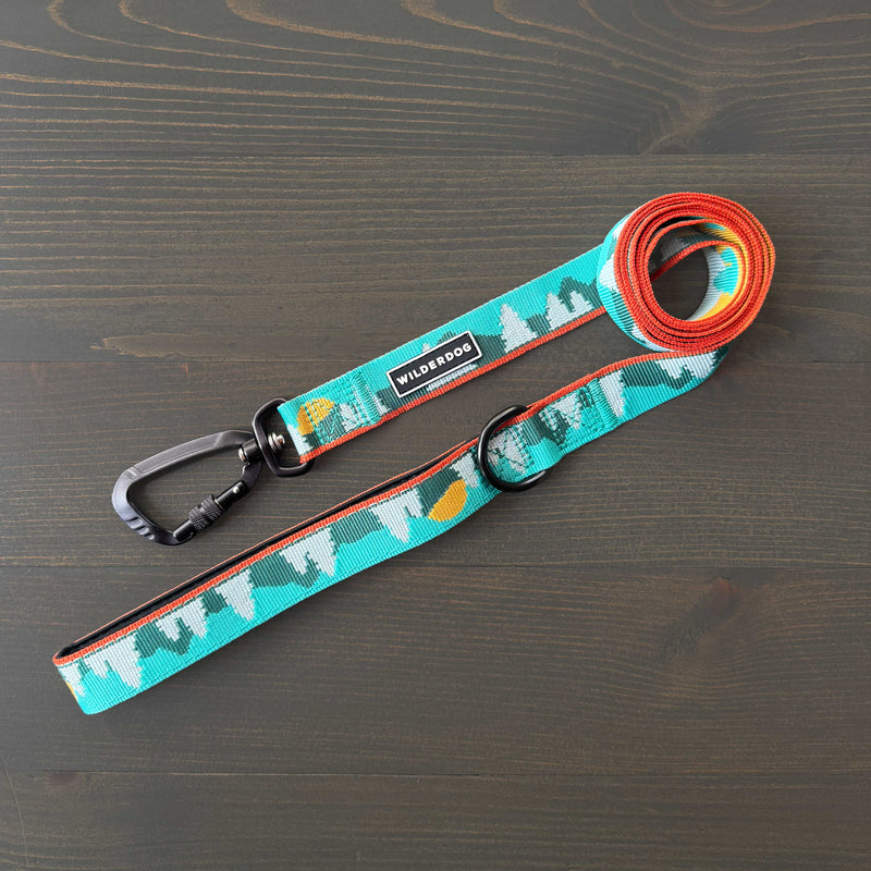 The Tamarack Ultralight Leash, a teal nylon leash with a mountain and sun pattern, metal clip, and black ring, is laid flat on a dark wooden surface. The “WILDERDOG” brand name is visible on the leash.