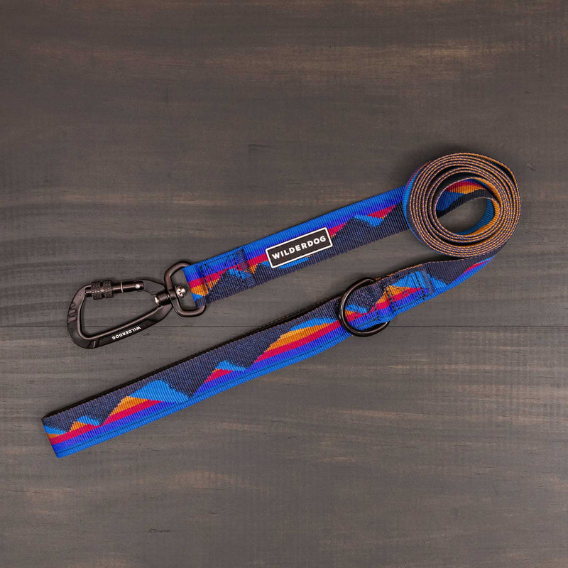 The Cordillera Ultralight Leash: a coiled blue leash with red, orange, and purple mountain patterns, ultralight nylon webbing, swiveling carabiner attachment, and a "Wilderdog" logo on a black label—shown on a dark wood surface.