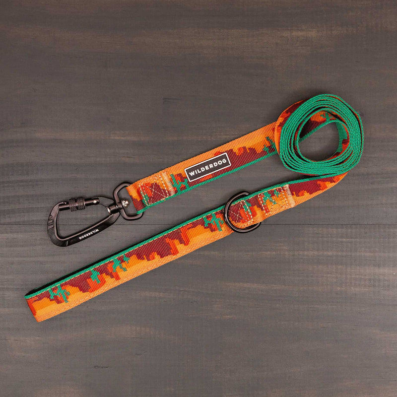 The Moab Ultralight Leash, featuring an orange, green, and red pattern, durable nylon webbing, a black "Wilderdog" label, and a metal clip, is laid out on a dark wooden surface.