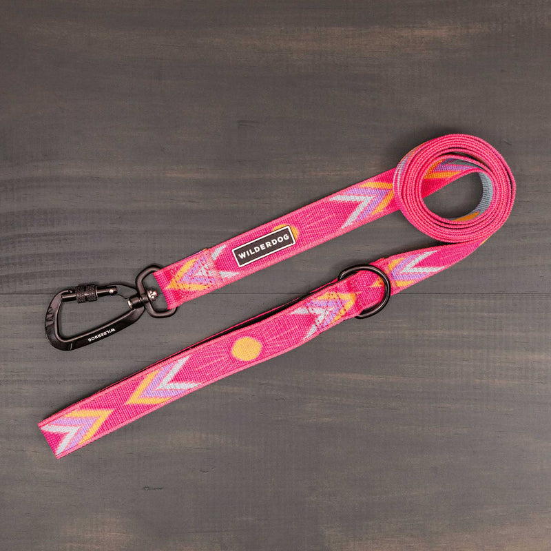 The Playa Ultralight Leash features pink nylon webbing with colorful zigzag patterns, a black metal clip and ring, and a "Wilderdog" label near the clip. Shown laid flat on a dark wooden surface.