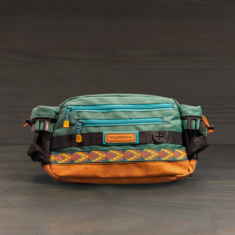 The Mega Hip Pack features teal and brown fabric, a patterned band, two front zippers, mesh side pockets, and a "WILDPROS" patch. This vegan hip pack is perfect for stylish, hands-free dog walking. Shown on a plain dark background.