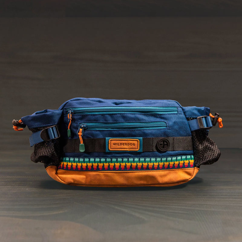 The Mega Hip Pack, shown in blue and tan with a geometric pattern, mesh side pockets, orange zipper pulls, and a "Wilderdog" patch on the front, is your ideal hands-free dog walking bag against a dark background.