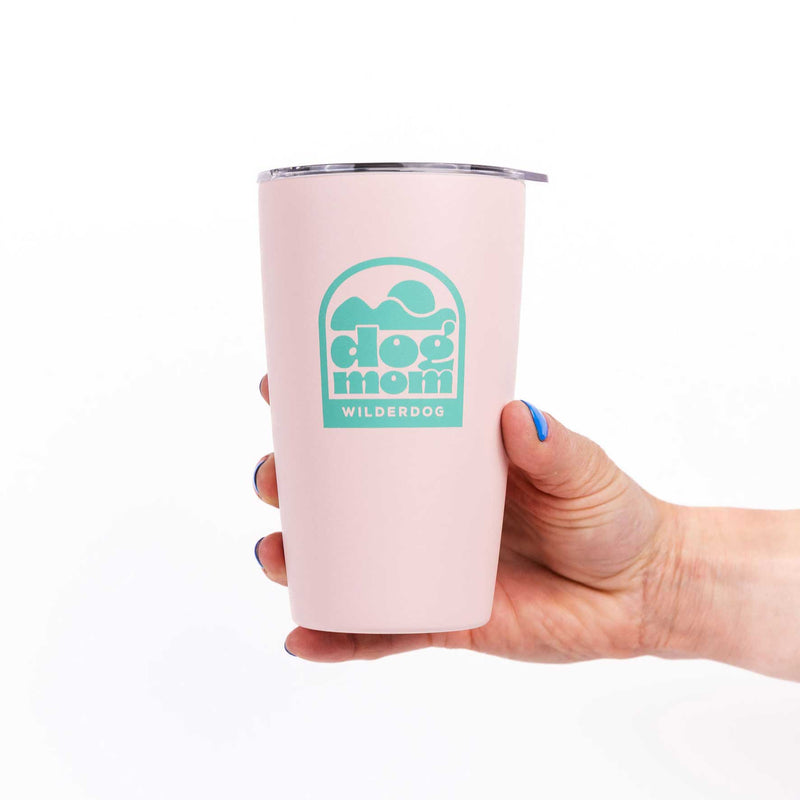 A hand with blue-painted nails holds the Dog Mom Mug, a light pink travel tumbler with a teal "dog mom" logo and "WILDERDOG" below; its stainless steel, double-wall insulation keeps drinks at the perfect temperature.