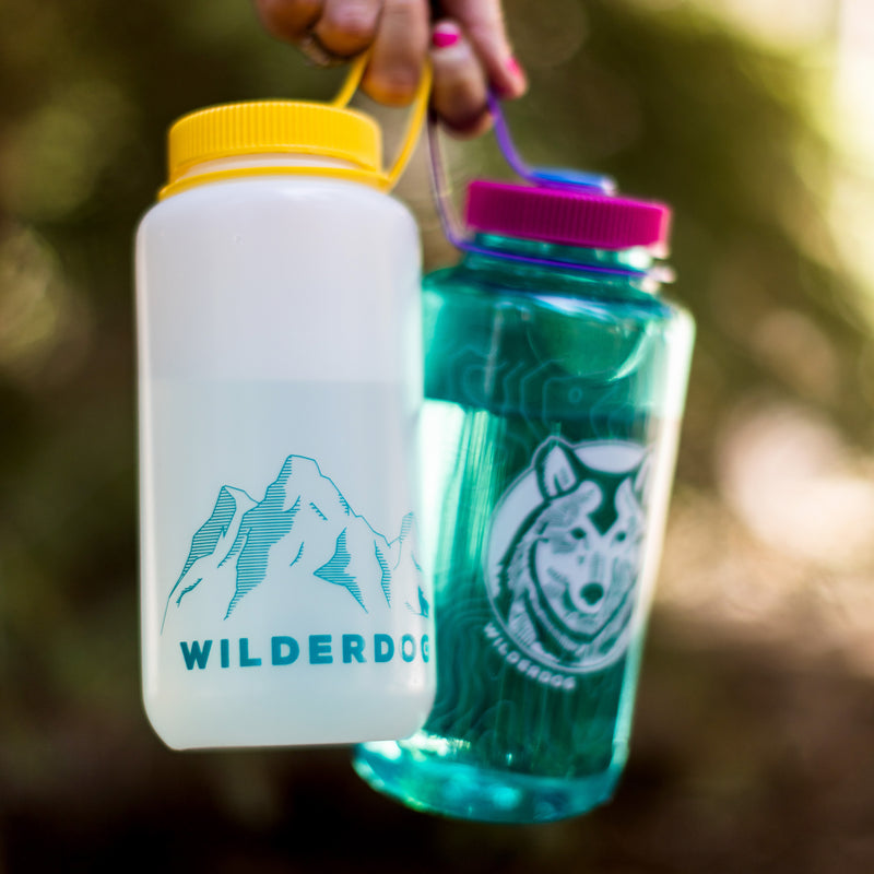 A person holding two colorful BPA/BPS-free Wilderdog Nalgene water bottles; one is a 32oz Wide Mouth, white with a yellow cap, featuring a mountain graphic and the text "WILDERDOG." The other is teal with a purple cap, depicting a dog's face in a circle with the words "WILDERDOG" around it.