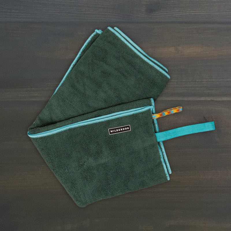 The Quick Dry Towel, dark green with turquoise trim and a "WILDSOOG" tag, lies folded on a dark wood surface. Two colorful hanging loops are attached to one corner, making it ideal for fast drying your dog.
