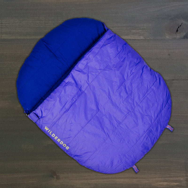 A Sleeping Bag in purple and blue is laid out flat on dark wood, ideal for your dog to use as a camping bed.