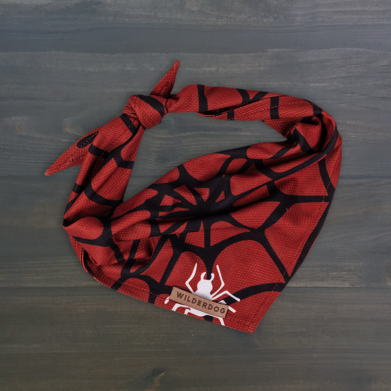The Spider Bandana is red and black with a web pattern, a white spider graphic, and a WilderDog tag on grey wood.