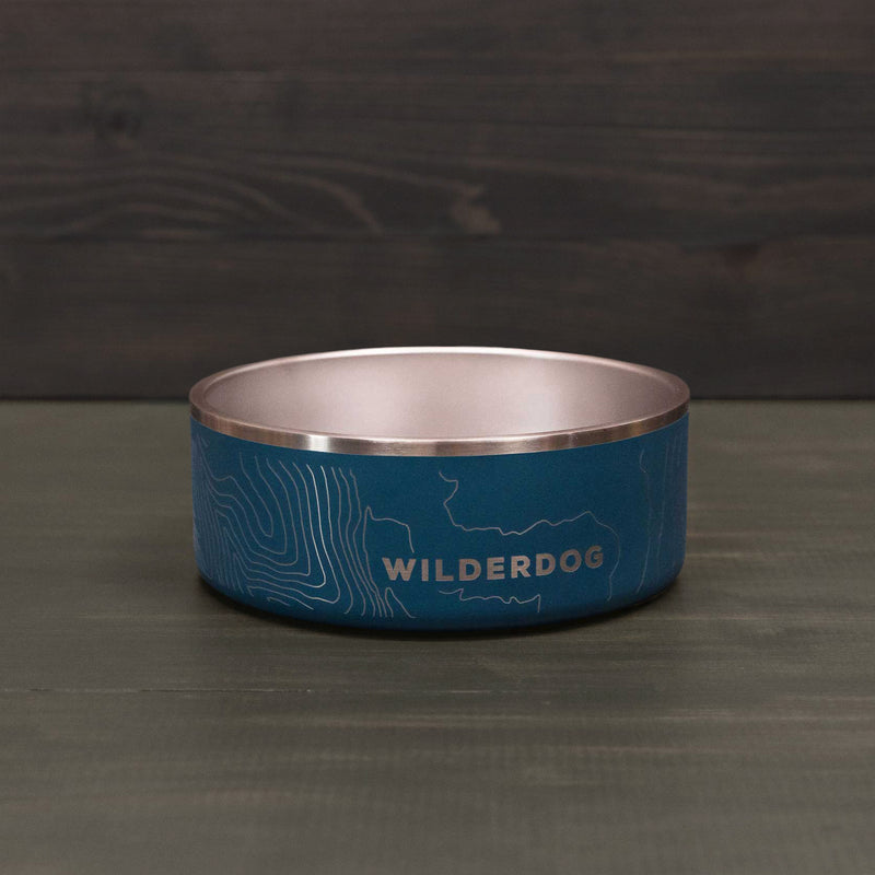 The Stainless Steel Dog Bowl, featuring a blue topographic map design and "WILDERDOG" printed on the side, sits on a dark wooden surface with a matching background.