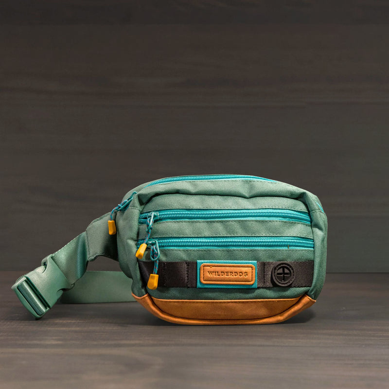 The Utility Pack is a small green and brown fanny pack with multiple zippered pockets, yellow zipper pulls, and a "WILDERDOG" patch on the front—ideal as a treat bag—shown on a wooden surface with a dark background.