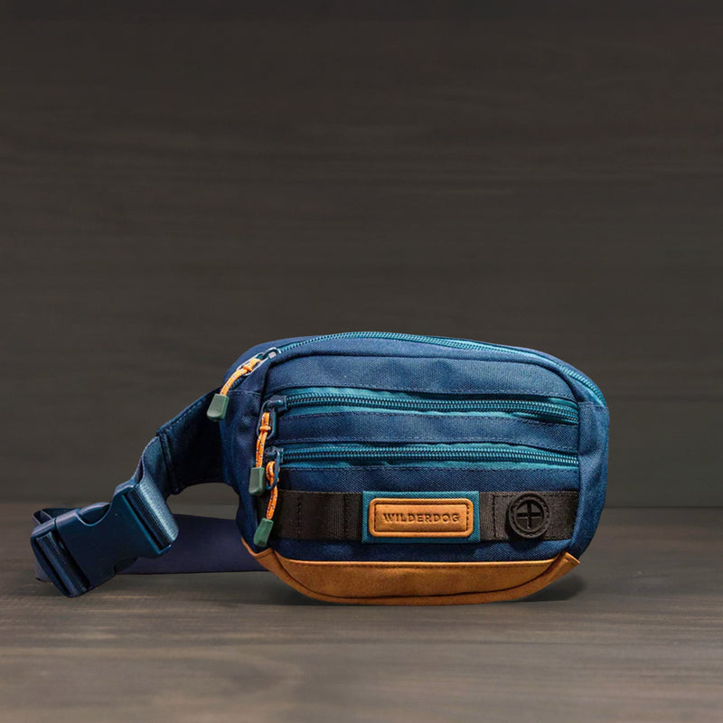 The Utility Pack is a small blue and brown fanny pack with several zippered pockets, a black strap, and a "Wilderdog" patch on the front, shown against a dark background.