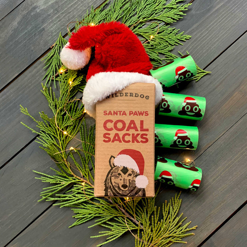 A box of Santa Paws Poop Bags, featuring a wolf in a Santa hat, is surrounded by festive poop bags decorated with Santa hats, string lights, and evergreens on a dark wooden surface.