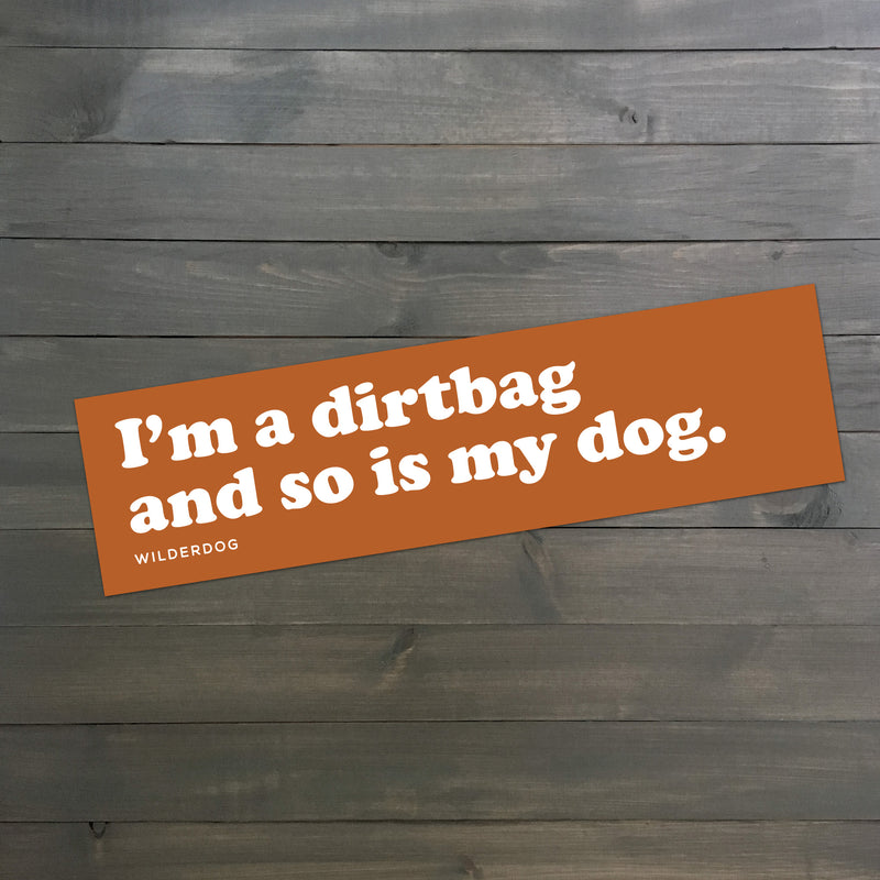 A rectangular orange bumper sticker with white text reading, "I'm a dirtbag and so is my dog," lies on a wooden surface with a grey finish. The word "WILDERDOG" is printed in smaller white text at the bottom left, making the Dirtbag Bumper Sticker perfect for any dog lover's car accessory collection.