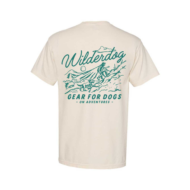The back of the Wilderdog Pocket Tee, a cream-colored unisex shirt, showcases teal graphics and text that say "Wilderdog" and "Gear for Dogs on Adventures." The design depicts a person hiking with their dog across a mountainous landscape, accented by an elegant wolf logo—ideal for individuals who appreciate sustainably produced clothing.