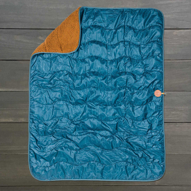 A blue quilted Picnic Blanket with rounded corners is laid flat on a dark wood surface. One corner folds over to reveal a brown, soft fleece-like lining. Enjoy comfort and peace of mind with our lifetime warranty on this cozy blanket.