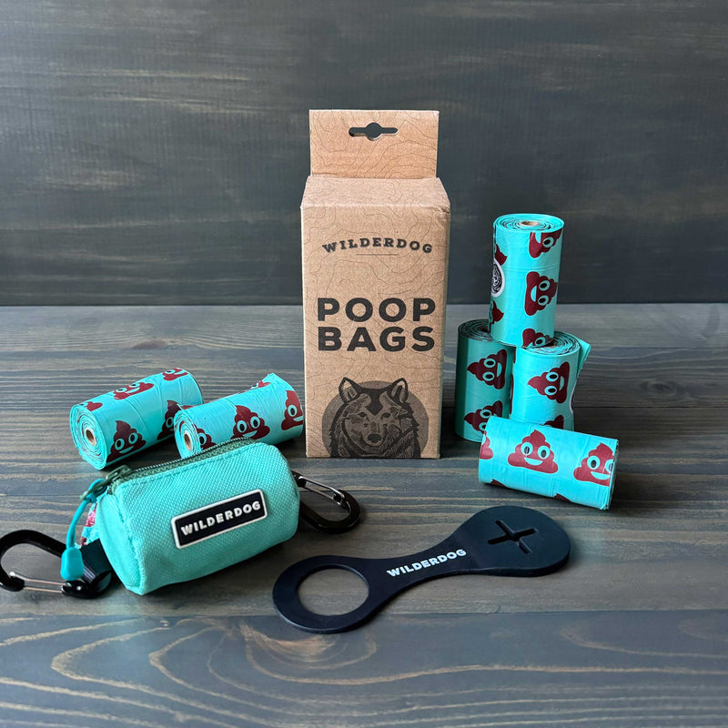 The Ultimate Poop Kit includes rolls of turquoise poop bags with cartoon poop emojis, a matching turquoise holder, and a black dispenser tool, all displayed on a wooden surface.