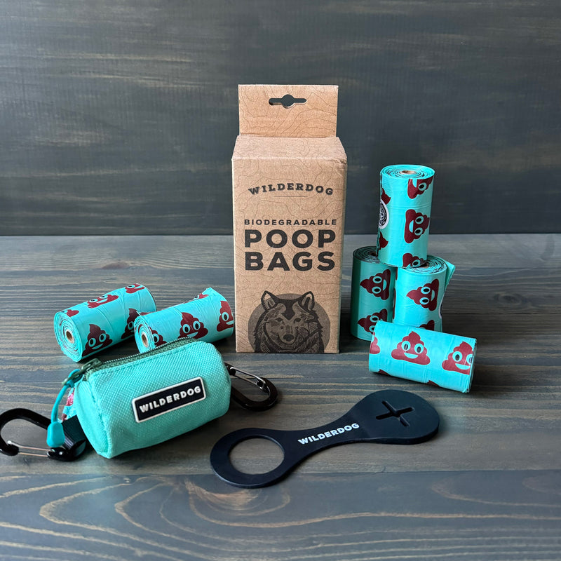 The Ultimate Poop Kit, which includes biodegradable poop bags decorated with cartoon poop emojis, is arranged on a table. The kit features a brown box, multiple rolls of bags, a turquoise carrying pouch with a clip, and a black dog poop holder dispenser, all branded by Wilderdog.