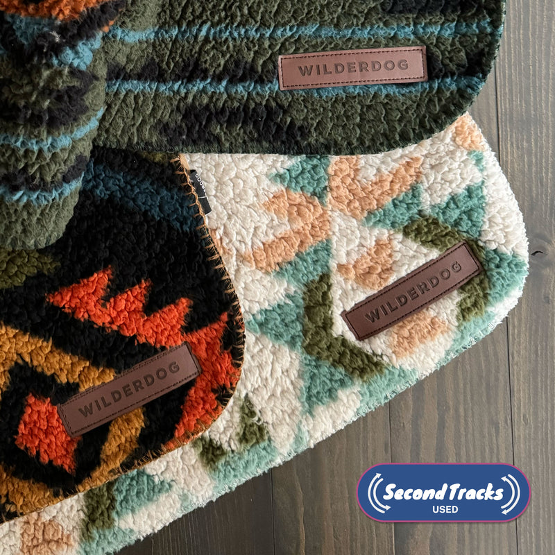 Three (Used) - Sherpa Blankets with "Wilderdog" labels, each featuring cozy fleece and waterproof lining, are layered on a dark wood surface; the "Second Tracks USED" logo is in the lower right corner.