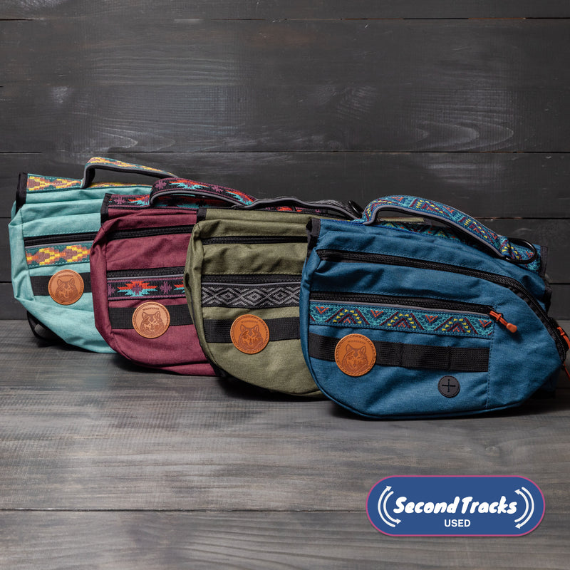 Four colorful (Used) Backpacks with decorative patterns and leather patches, including a mystery color, are lined up on a wooden surface. A “Second Tracks USED” logo appears in the lower right corner.