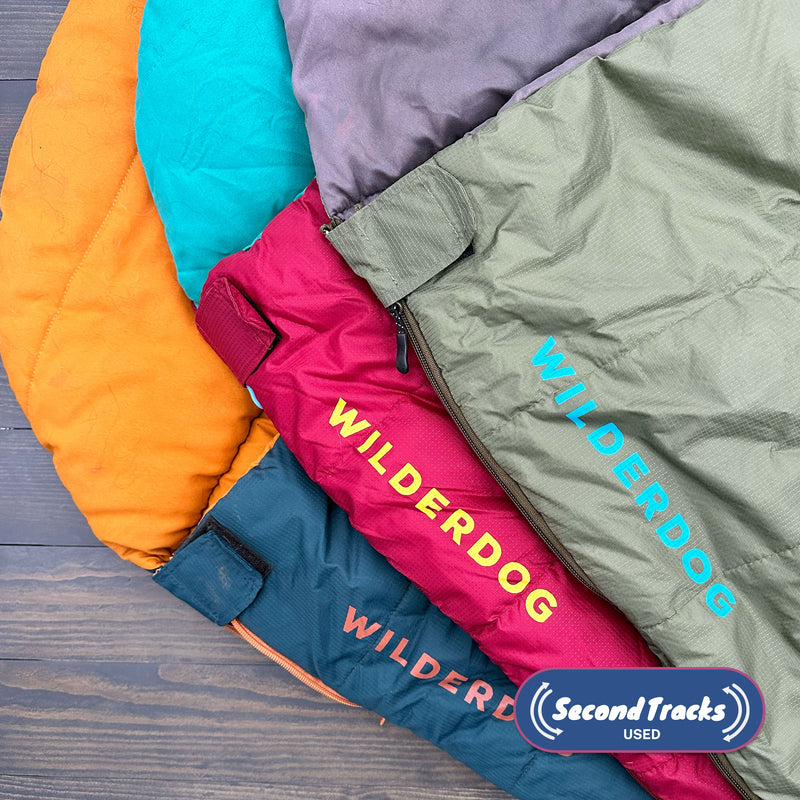 Four colorful (Used) Sleeping Bags—orange, teal, magenta, and olive—are stacked on wood. Durable and perfect for outdoor adventures, each shows a "SecondTracks USED" logo in the bottom right corner.