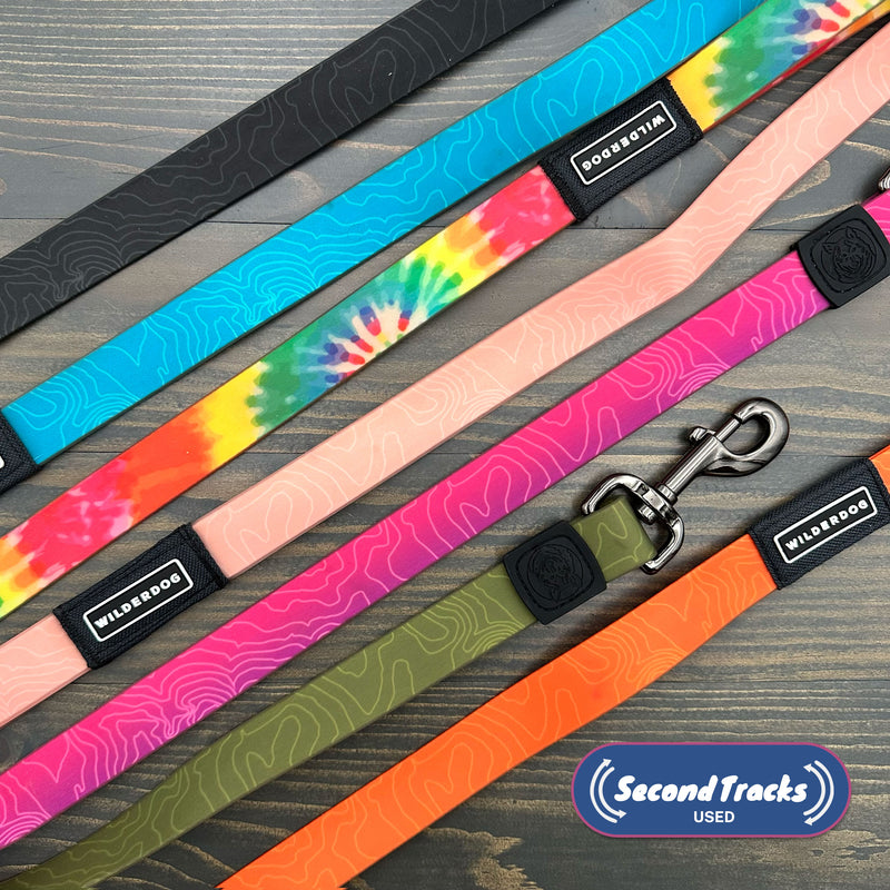 Five colorful dog leashes, including a Mystery Color Leash and the (Used) Waterproof Leash, are arranged diagonally on a wooden surface. Designs feature tie-dye and topographic lines. "Second Tracks Used" logo appears in the lower right.