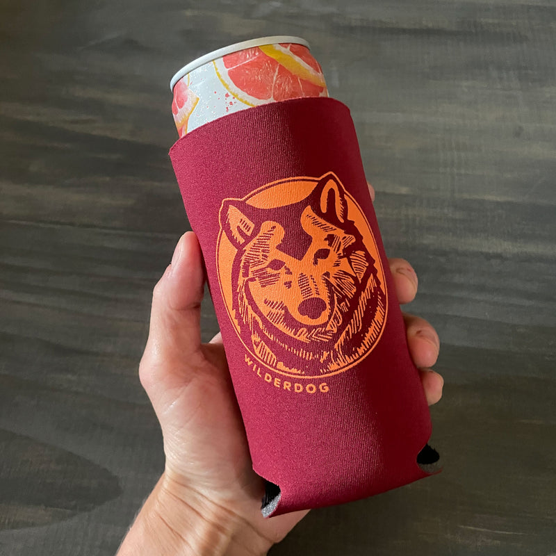 A hand holds a skinny can wrapped in a red "Slim Can Wolf Koozies," featuring an orange illustration of a wolf's head and the word "Wilderdog" below it. The background is a dark, textured surface, making this one of those Party Dog Koozies you'll love.