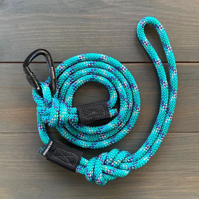 The Islander Reflective Small Carabiner Leash, showcasing a coiled turquoise rope with interwoven blue, white, and purple patterns, lies on a wooden surface. Crafted from durable rock climbing rope, it features a black carabiner clip on one end and a black fabric loop on the other end, ensuring both reliability and sturdiness for any adventure.