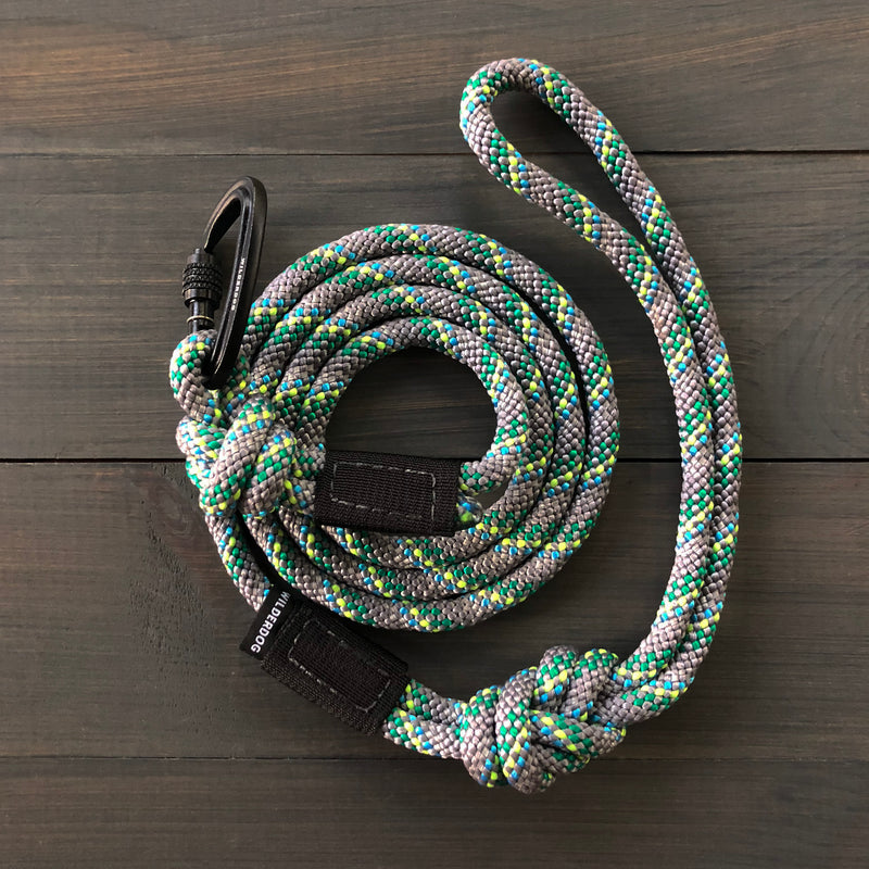 An Alpine Reflective Small Carabiner Leash, featuring coiled multicolored patterns of green, blue, and gray, lies on a dark wooden surface. The durable leash is attached to a black carabiner clip at one end, with a black strip of fabric and a small tag affixed near the clip.