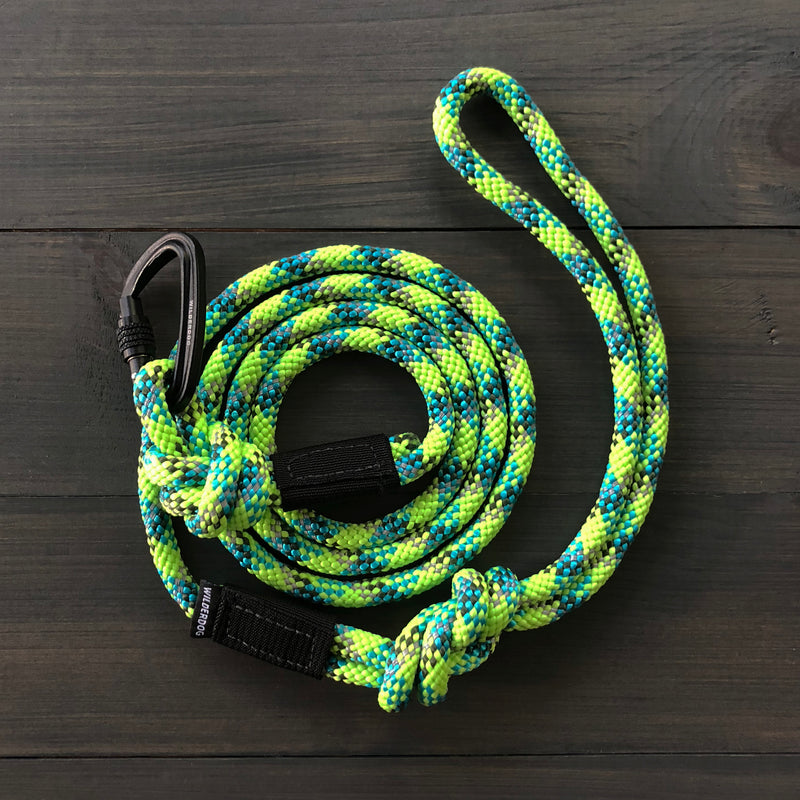 The Lime Reflective Small Carabiner Leash, made from durable rock climbing rope in a vibrant blue, green, and yellow pattern, lies coiled on a dark wooden surface. The reflective leash features a loop handle on one end and has a black clasp attached with a knot near the clasp.