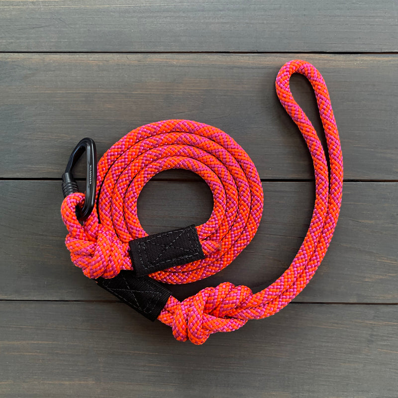 The Sierra Small Carabiner Leash, made from robust rock climbing rope and featuring a vibrant pink and orange coiled design, is laid out on a wooden surface. The durable black metal clip is securely attached and reinforced with black fabric at the base, while a convenient loop handle sits at one end.