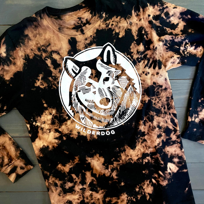 The Reverse Dye Long Sleeve Wolf Shirt is a hand-dyed black and beige long-sleeve shirt featuring a line-drawn image of a wolf's head in the center within a circular border. Below the image, "WILDERDOG" is printed in white capital letters. This unisex shirt effortlessly combines style and versatility.
