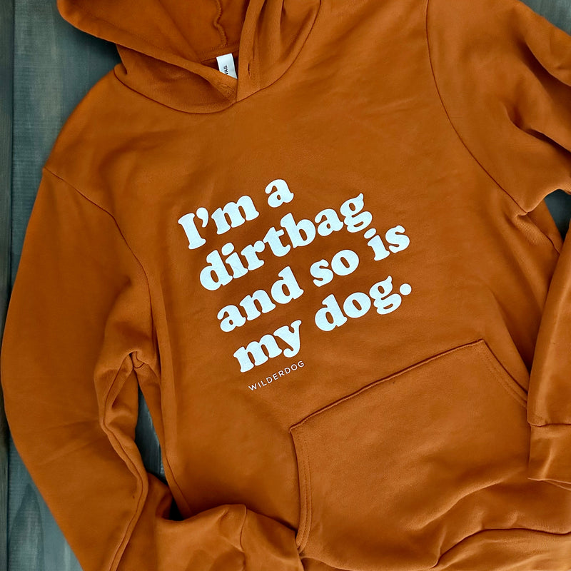 A Dirtbag Hoodie, unisex and brown with a front pocket, lays flat on a dark surface. Made from a soft cotton/poly blend, it features white text that reads, "I'm a dirtbag and so is my dog," with "Wilderdog" in smaller letters below. It's the softest sweatshirt you'll ever wear.