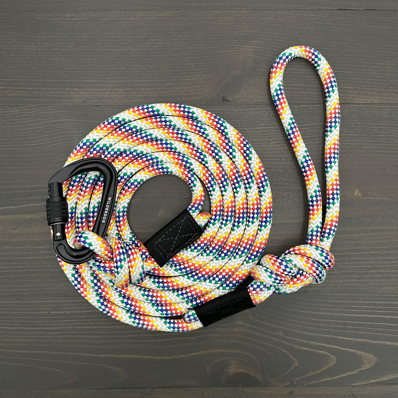The Rainbow Leash, a coiled, multi-colored braided rope with a black carabiner at one end, lies on a wooden surface. The other end is looped and secured with a knot—perfect as a versatile dog leash or durable rock climbing rope that comes with a lifetime warranty.