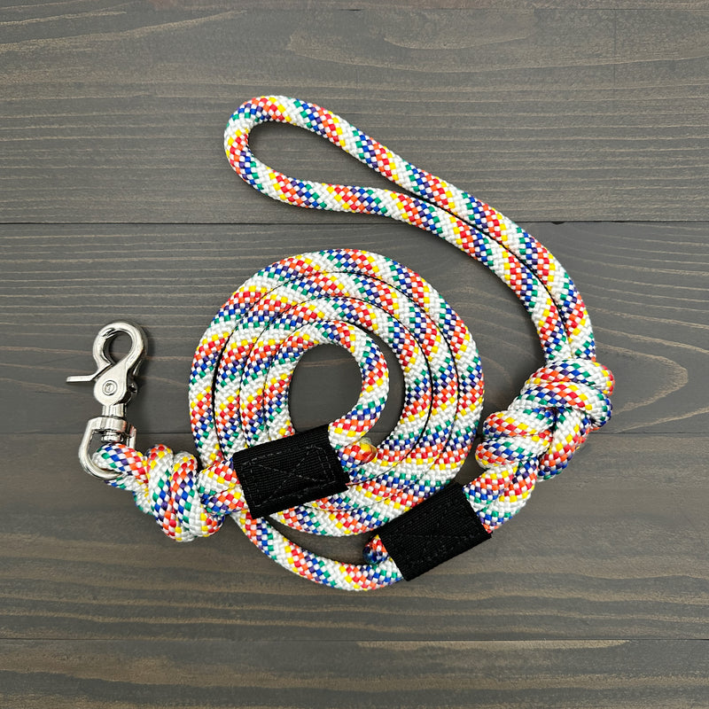 The Rainbow Quick Clip Leash, a colorful and durable leash crafted from woven, multicolored rock climbing rope, is coiled neatly on a dark wooden surface. This quick clip leash features a metal clasp at one end and a loop handle at the other.