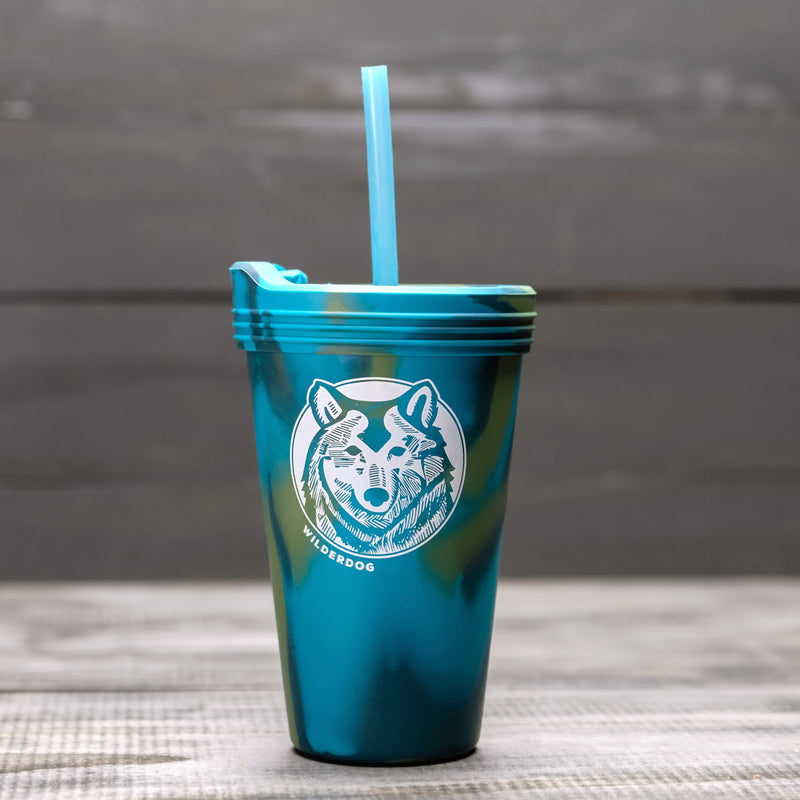 The 16 oz Silipint Pint Glass is a metallic blue travel cup featuring an illustration of a wolf's face, with the word "WILDERDOG" beneath it. It comes with a matching straw and has a spill-resistant silicone lid for added convenience. The background showcases a blurred wooden surface.
