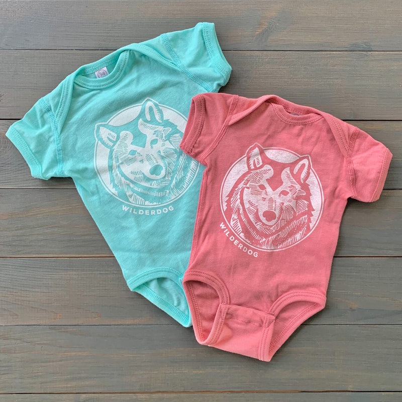 Two Wilderbaby Onesies rest on a wooden surface. The unisex baby onesie on the left is turquoise with a white graphic of a dog's face and the word "WILDERDOG." The furless puppy onesie on the right is pink with the same graphic and text. Both have short sleeves and round necks.